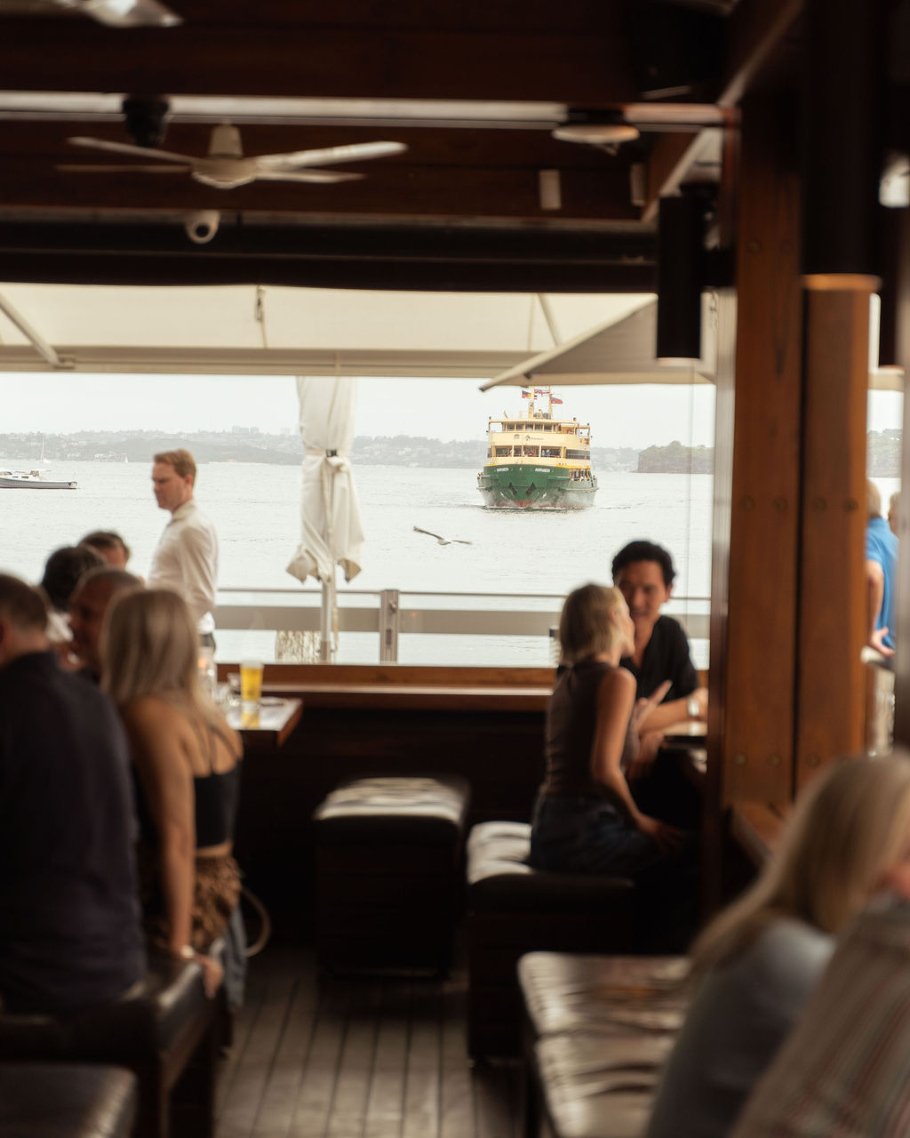 Guests in the Hugos Manly cocktail lounge as a Manly ferry passes the window