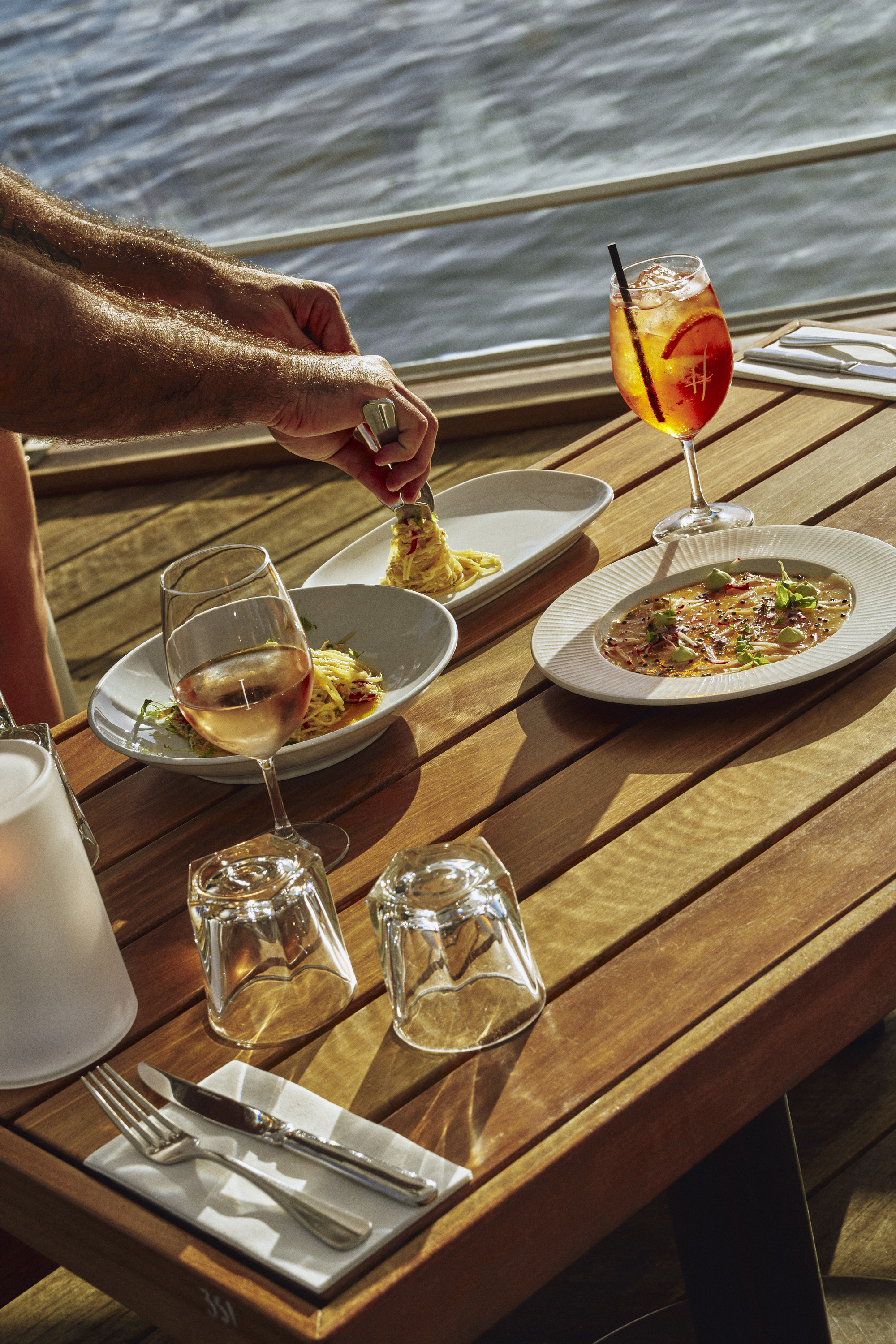 Plated main courses and a spritz on a timber table beside the water at Hugos Manly